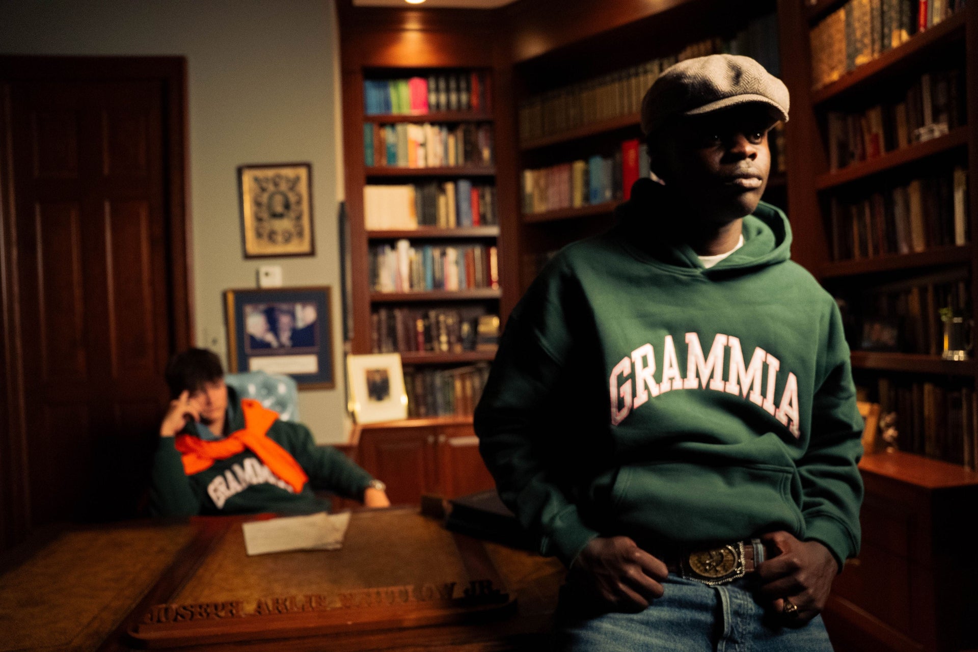 A man in a flat cap leans on a desk in a wood-panelled library, wearing the green Grammia hoodie. A second man sits behind him with an orange sweater over his shoulders.
